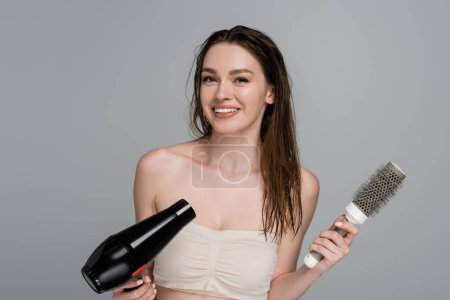 Photo for Cheerful young woman with wet hair holding round hair brush and hair dryer isolated on grey - Royalty Free Image