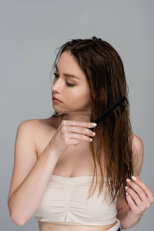 pretty young woman with bare shoulders combing wet hair isolated on grey 