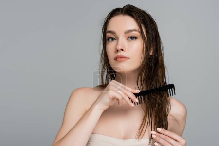 Téléchargez les photos : Jolie jeune femme aux épaules nues brossant les cheveux mouillés avec peigne isolé sur gris - en image libre de droit