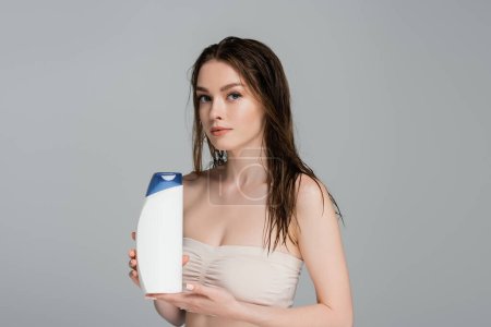 Téléchargez les photos : Jolie femme aux cheveux mouillés et épaules nues tenant bouteille de shampooing isolé sur gris - en image libre de droit