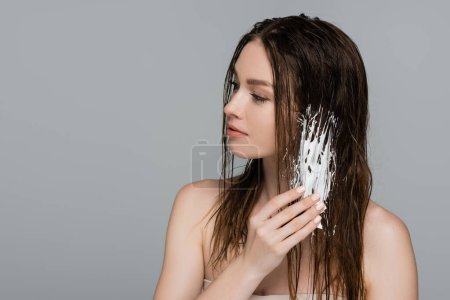 hübsche junge Frau mit nackten Schultern, die Schaum auf das nasse Haar aufträgt, isoliert auf grau