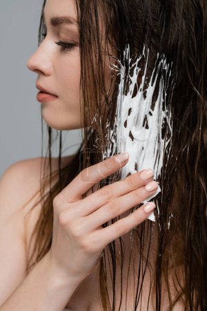 pretty young woman with bare shoulder applying foam while styling wet hair isolated on grey