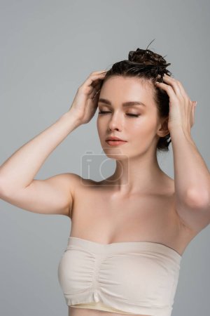 bonita mujer joven con los ojos cerrados lavando el cabello aislado en gris 