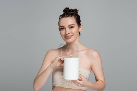 cheerful young woman with wet hair holding container with mask isolated on grey 