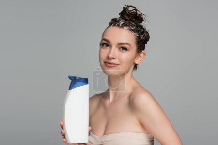 fröhliche junge Frau mit nassen schäumenden Haaren, die Flasche mit Shampoo isoliert auf grau hält 