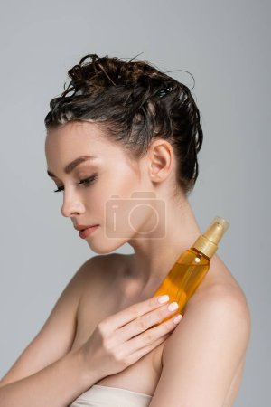 brünette junge Frau mit nassen Haaren hält Flasche mit Behandlungsöl isoliert auf grau 