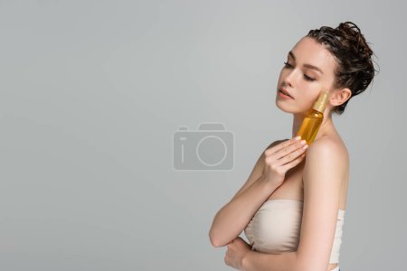 Photo for Brunette young woman with wet hair holding bottle with oil isolated on grey - Royalty Free Image