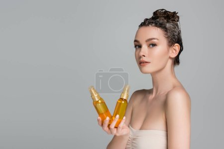 brunette jeune femme aux cheveux mouillés tenant des bouteilles avec de l'huile de traitement isolé sur gris 