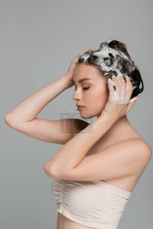 young woman with closed eyes washing wet and foamy hair isolated on grey 
