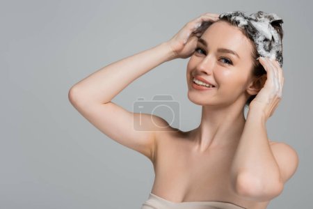 fröhliche junge Frau wäscht nasses und schäumendes Haar isoliert auf grau