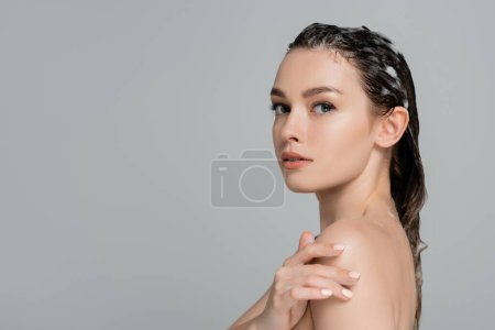 junge Frau mit schäumenden und nassen Haaren, die die nackte Schulter berührt, isoliert auf grau 