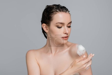junge Frau mit nassen Haaren berührt nackte Schulter mit weißem Schaum isoliert auf grau 