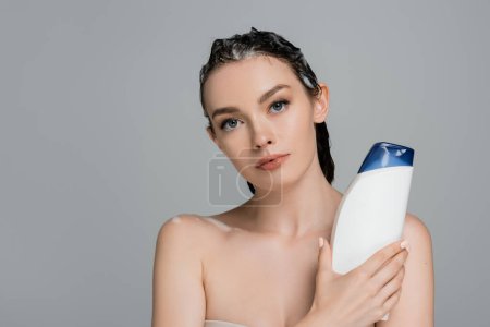 brünette junge Frau mit nassen schäumenden Haaren, Flasche mit Shampoo isoliert auf grau 