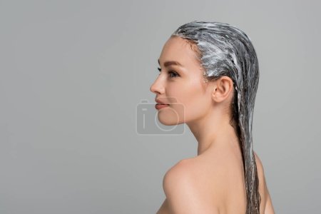 Téléchargez les photos : Vue latérale de la jeune femme joyeuse avec épaules nues et masque capillaire isolé sur gris - en image libre de droit