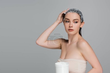 jeune femme aux épaules nues appliquant un masque sur cheveux mouillés et tenant un bocal isolé sur gris 