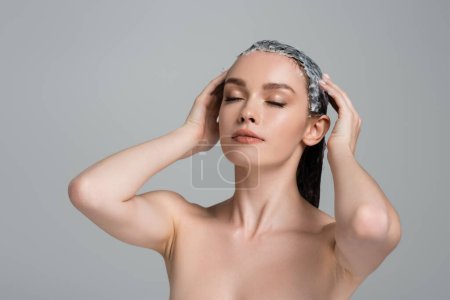 jeune femme aux yeux fermés appliquant masque sur cheveux mouillés isolé sur gris 