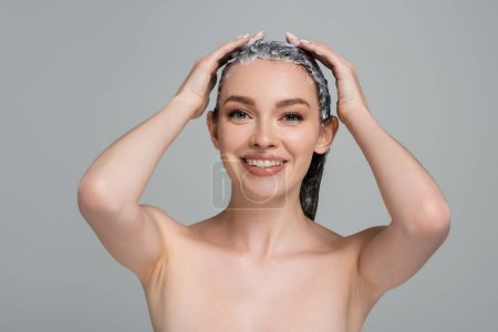mujer joven feliz con hombros desnudos aplicando máscara sobre el cabello mojado aislado en gris 