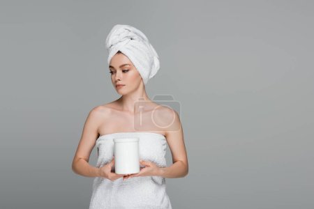 young woman with white towel on head holding jar with hair mask isolated on grey 