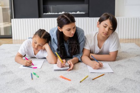 Asiatische Kinder zeichnen auf Papier in der Nähe der Mutter, die zu Hause auf Teppich liegt 