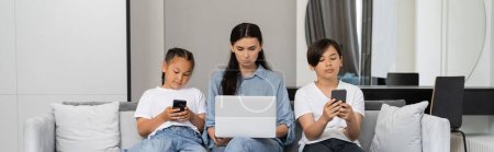 Photo for Mother working on laptop near asian kids with smartphones at home, banner - Royalty Free Image