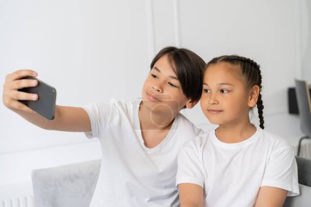 Foto de Preadolescente asiático chico tomando selfie en celular con hermana en casa - Imagen libre de derechos