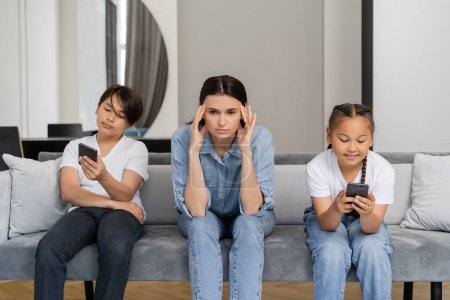 Woman suffering from headache near asian kids using smartphones at home 