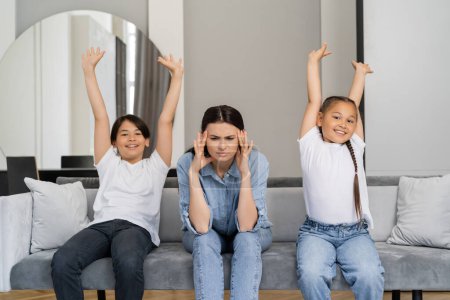 Cheerful asian kids sitting with raised hands near tired mother on couch at home 
