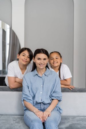 Lächelnde asiatische Kinder schauen in die Kamera nahe der Mutter auf der heimischen Couch 