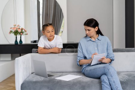 Asian kid looking at mom holding notebook near laptop at home 