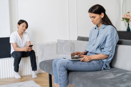 Photo for Woman using laptop near blurred asian son with smartphone at home - Royalty Free Image