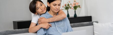 Photo for Asian kid hugging mom looking at laptop on couch at home, banner - Royalty Free Image