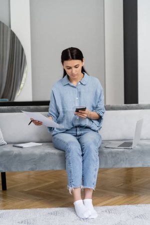 Foto de Freelancer en camisa usando celular y sujetando papel cerca del portátil en el sofá en casa - Imagen libre de derechos