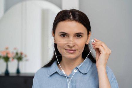 Portrait of brunette woman using earphones and looking at camera at home 
