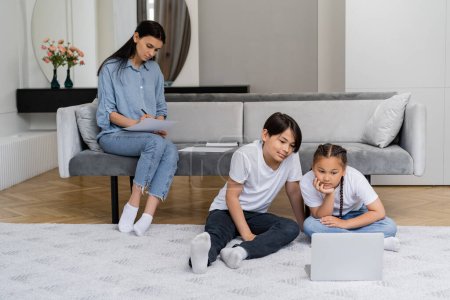 Asiático niños mirando portátil cerca de madre escritura en papel en sala de estar 