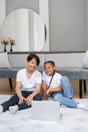 Cheerful asian siblings watching film on laptop on floor at home 