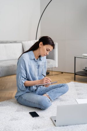 Brünette Frau schreibt auf Notizbuch in der Nähe von Geräten auf Teppich zu Hause 