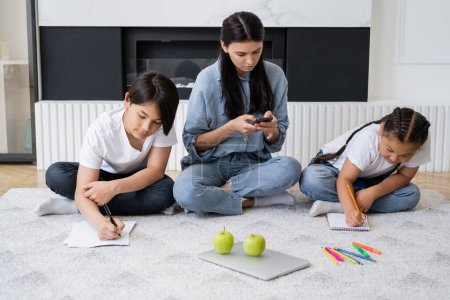 Umtriebige Mutter benutzt Smartphone, während asiatische Kinder zeichnen und zu Hause auf dem Boden sitzen 