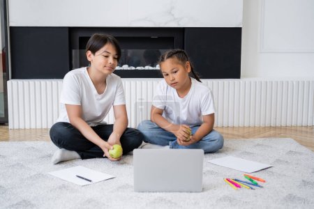 Téléchargez les photos : Les enfants asiatiques tenant des pommes et regardant un film sur ordinateur portable près du papier et des crayons de couleur à la maison - en image libre de droit