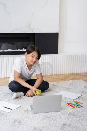 Preteen asian boy holding apple near laptop and color pencils at home 