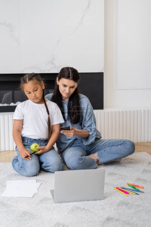 Mother using smartphone and laptop near asian daughter with apple and color pencils at home 