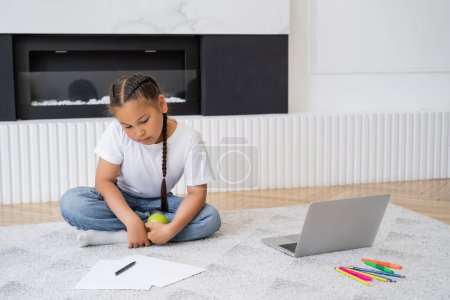 Asian girl holding apple near laptop and color pencils at home 