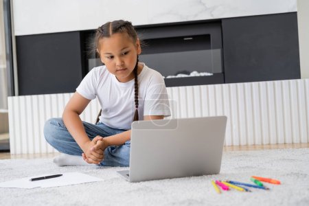 Niño asiático viendo dibujos animados en el ordenador portátil cerca de papeles y lápices de color en casa 