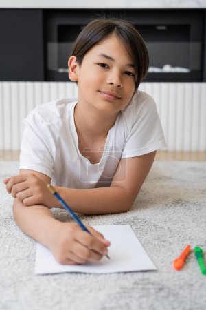 Preteen asian boy looking at camera while drawing on paper at home 