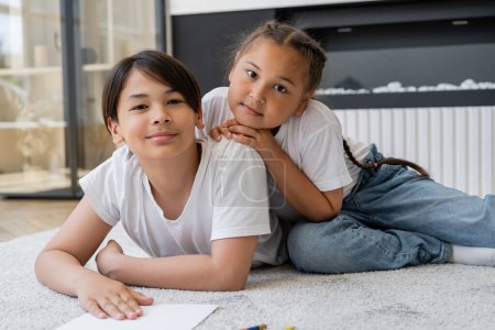 Asian siblings looking at camera while lying on floor near paper at home 