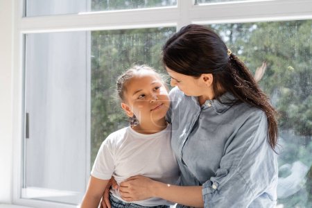 Frau umarmt asiatische Tochter am Fenster ihres Hauses 