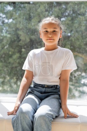 Portrait of asian girl looking at camera while sitting on windowsill at home 