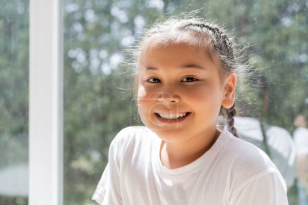 Portrait of cheerful asian girl looking at camera near window at home 