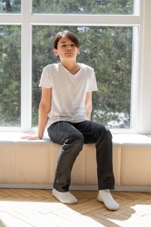Foto de Preadolescente asiático chico sentado en windowsill en casa - Imagen libre de derechos