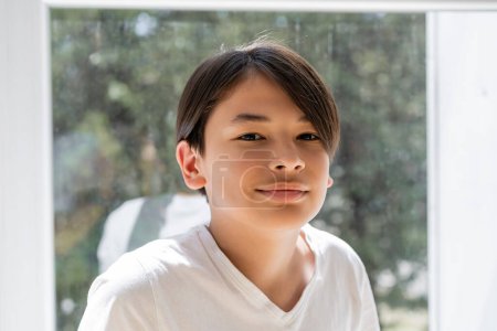 Retrato de preadolescente asiático chico sonriendo en cámara cerca de ventana en casa 