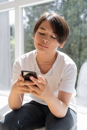 Preteen asian boy using smartphone while sitting on windowsill at home 